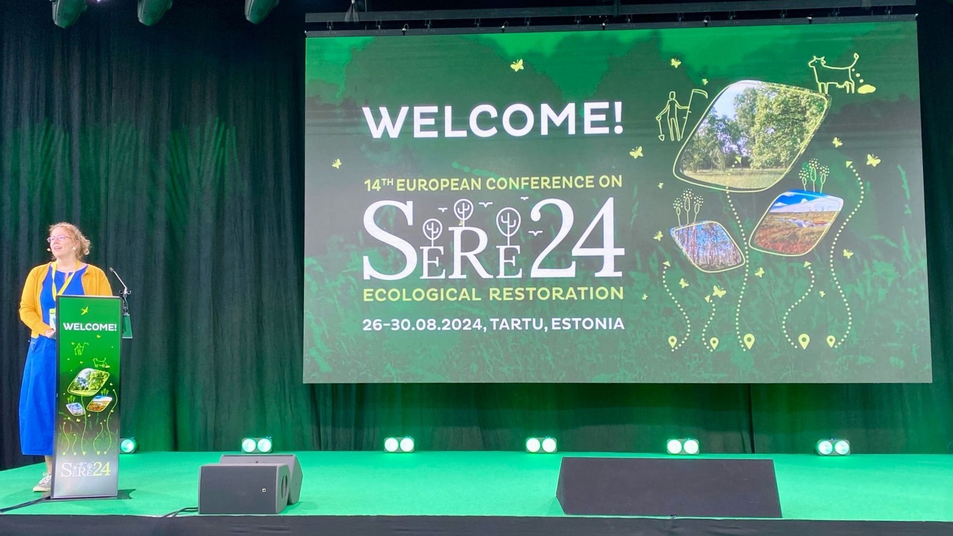 A woman in a blue dress and yellow cardigan stands at a podium on a stage with a large green screen behind her that says "WELCOME! 14th EUROPEAN CONFERENCE ON SERE24 ECOLOGICAL RESTORATION 26-30.08.2024, TARTU, ESTONIA" with illustrations of nature and wildlife.